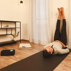 Mann in entspannter Pose mit Beinen an der Wand, Teil seiner yoga abendroutine. Eine Matte, ein Handtuch und eine Massageball-Box liegen bereit.