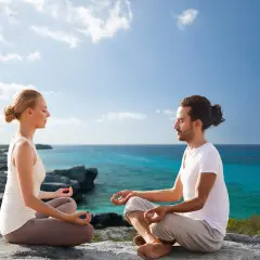 Ein Paar meditiert in Lotusposition an einer felsigen Küste. Sie suchen nach spirituellen Ausbildungen, um ihre innere Ruhe zu finden.