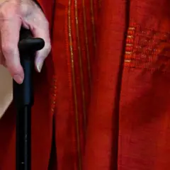 Hand eines Priesters im Ruhestand, der einen Gehstock hält. Er trägt eine rote Soutane mit goldenen Verzierungen.