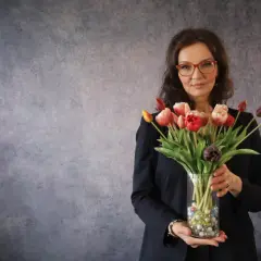Eine energetische Person mit Brille hält einen Strauß bunter Tulpen in einem Glasvase.