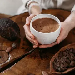 Hände halten eine Tasse heißen Kakao, umgeben von Kakaobohnen und einer Kakaoschote. Ein Moment der Ruhe bei einer Kakao-Zeremonie.