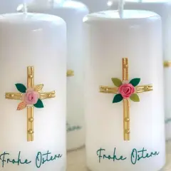 Mehrere weiße Osterkerzen mit goldenen Kreuzen, rosa Rosen und grünen Blättern. Die Osterkerze Bedeutung wird durch diese Symbole unterstrichen. "Frohe Ostern" steht geschrieben.