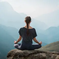 Frau in Hatha Yoga Pose auf einem Felsen sitzend, mit Blick auf eine neblige Berglandschaft.