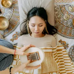 Eine Frau liegt entspannt, während eine andere Person ihr eine Kalimba über dem Bauch hält. Klangschalen und ein Stößel sind um sie herum arrangiert, was auf eine Klang Meditation hindeutet.
