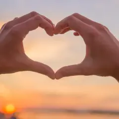 Hände formen ein Herz vor einem Sonnenuntergang. Eine spirituelle Person drückt Liebe und Verbundenheit aus.