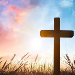 Ein Kreuz ragt im Abendrot auf, umgeben von Gräsern. Möge es ein gesegnetes Osterfest sein.