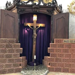 Ein Kreuz mit Jesusfigur, umgeben von Ziegelmauern und lila Vorhängen. Ein Kerzenlicht lädt zum Kreuz Gebet ein.