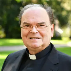 Bischof Bertram lächelt in die Kamera, trägt ein schwarzes Gewand mit weißem Kragen und eine Brille. Im Hintergrund ist ein grüner Park zu sehen.
