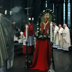 Ein Kardinal leitet eine gesungene katholische Messe. Ein Bildnis des Papstes, umrahmt von Blumen, steht im Mittelpunkt.