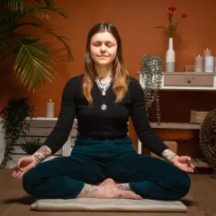 Frau in tiefer meta meditation, umgeben von Pflanzen und Kerzen. Eine Shiva-Statue im Hintergrund.