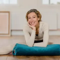 Frau lächelt in einem hellen Raum, Beine gespreizt, mit einem blauen Bolster für Nadi Yoga.