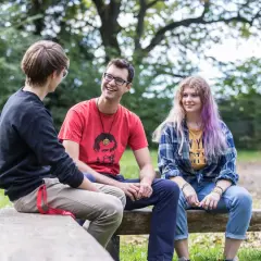 Drei junge Leute unterhalten sich lachend auf einer Bank im Freien. Der Mann in Rot trägt ein T-Shirt mit einem Porträt, das an den **Salesianer Orden** erinnert.