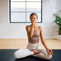 Frau praktiziert Pranayama Atmung im Lotussitz auf einer Yogamatte. Sie hält ihren Finger vor die Nase, um abwechselnd durch die Nasenlöcher zu atmen.