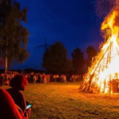 Menschen versammeln sich, um die Sommersonnenwende zu feiern. Ein riesiger Holzstapel brennt lichterloh im Abendhimmel, Funken steigen auf.