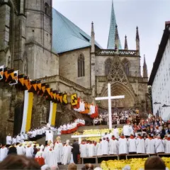 Große Menschenmenge vor einer Kathedrale, die an einem Hochamt teilnimmt. Fahnen und Girlanden schmücken die Fassade.
