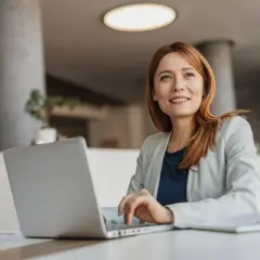 Eine Frau mit roten Haaren arbeitet an ihrem Laptop. Sie lächelt und blickt nach oben, als ob sie über ihre nächste Coaching-Sitzung nachdenkt. Ihr life coach job erfüllt sie.