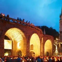 Eine Lichterprozession in Lourdes. Menschen halten Kerzen unter einem steinernen Bogen, während im Hintergrund eine Kirche beleuchtet ist.