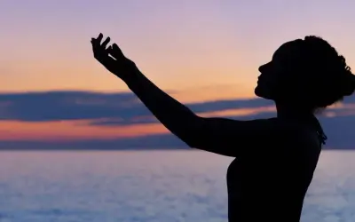 Silhouette einer Frau mit erhobenen Armen vor einem farbenfrohen Himmel und Meer. Sie scheint einen **chant deutsch** zu singen.