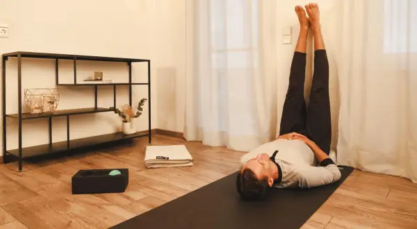 Mann in entspannter Pose mit Beinen an der Wand, Teil seiner yoga abendroutine. Eine Matte, ein Handtuch und eine Massageball-Box liegen bereit.