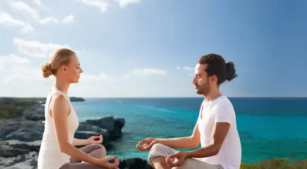 Ein Paar meditiert in Lotusposition an einer felsigen Küste. Sie suchen nach spirituellen Ausbildungen, um ihre innere Ruhe zu finden.