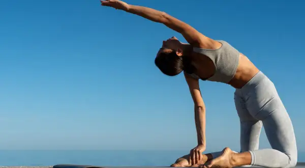 Frau in Yoga-Pose, ideal für Yoga Einsteiger, dehnt sich am Meer unter blauem Himmel.