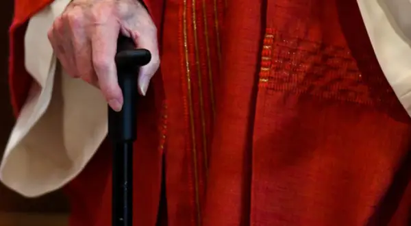 Hand eines Priesters im Ruhestand, der einen Gehstock hält. Er trägt eine rote Soutane mit goldenen Verzierungen.