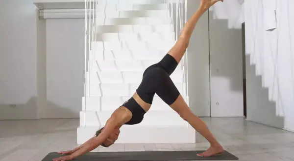 Frau in schwarzer Sportkleidung praktiziert Jivamukti Yoga in der Hundepose mit einem Bein erhoben, vor einer modernen weißen Treppe.