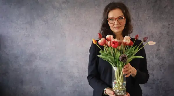 Eine energetische Person mit Brille hält einen Strauß bunter Tulpen in einem Glasvase.