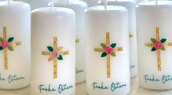 Mehrere weiße Osterkerzen mit goldenen Kreuzen, rosa Rosen und grünen Blättern. Die Osterkerze Bedeutung wird durch diese Symbole unterstrichen. "Frohe Ostern" steht geschrieben.