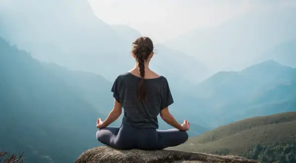 Frau in Hatha Yoga Pose auf einem Felsen sitzend, mit Blick auf eine neblige Berglandschaft.