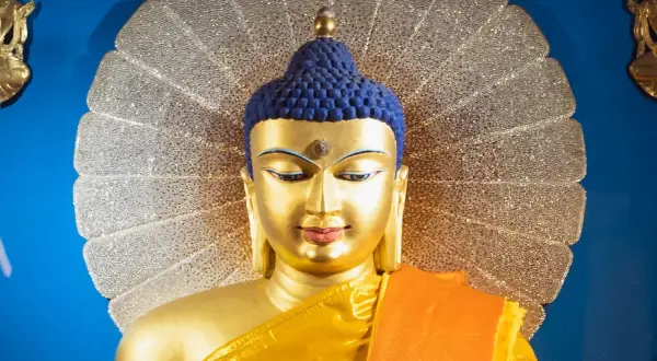 Goldene Buddha-Statue mit blauem Haar und orangefarbenem Gewand vor einem schimmernden Heiligenschein. Ein Wegweiser, wie werde ich buddhist.