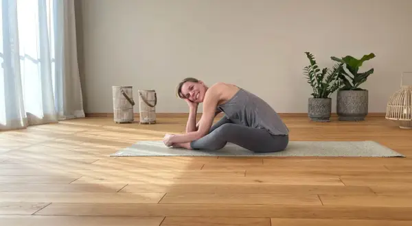 Frau in grauer Kleidung praktiziert Yoga Yin auf einer Matte. Sie lächelt entspannt und stützt ihren Kopf auf der Hand.