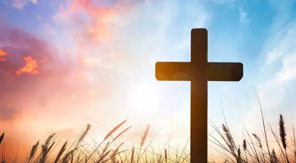 Ein Kreuz ragt im Abendrot auf, umgeben von Gräsern. Möge es ein gesegnetes Osterfest sein.