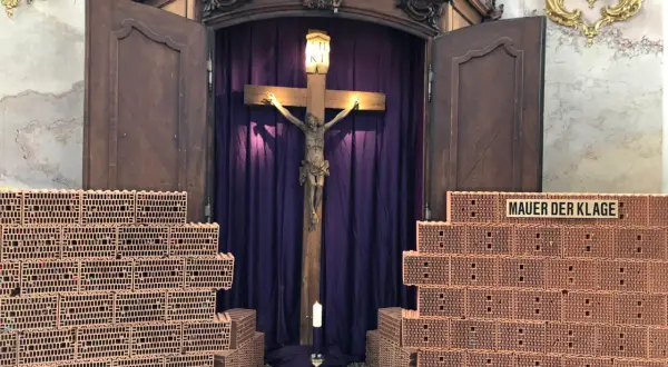 Ein Kreuz mit Jesusfigur, umgeben von Ziegelmauern und lila Vorhängen. Ein Kerzenlicht lädt zum Kreuz Gebet ein.