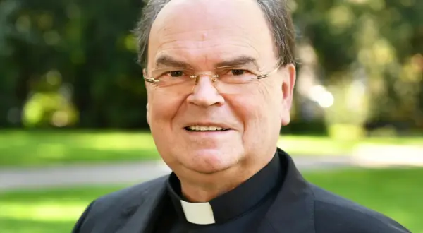 Bischof Bertram lächelt in die Kamera, trägt ein schwarzes Gewand mit weißem Kragen und eine Brille. Im Hintergrund ist ein grüner Park zu sehen.