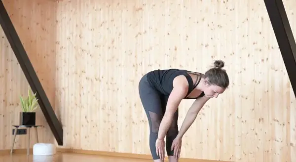 Frau in Uttanasana, einer Vorwärtsbeuge im Surya Namaskar Yoga, dehnt sich auf einer Matte.