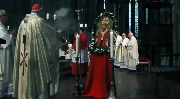 Ein Kardinal leitet eine gesungene katholische Messe. Ein Bildnis des Papstes, umrahmt von Blumen, steht im Mittelpunkt.