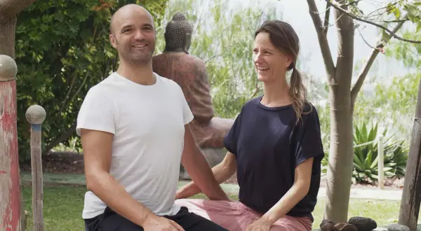 Ein Mann und eine Frau lächeln in einer grünen Umgebung. Sie sind die Gastgeber eines Ayurveda-Podcasts, der sich mit Wohlbefinden und Balance beschäftigt.