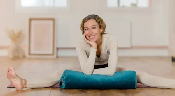 Frau lächelt in einem hellen Raum, Beine gespreizt, mit einem blauen Bolster für Nadi Yoga.