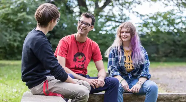 Drei junge Leute unterhalten sich lachend auf einer Bank im Freien. Der Mann in Rot trägt ein T-Shirt mit einem Porträt, das an den **Salesianer Orden** erinnert.
