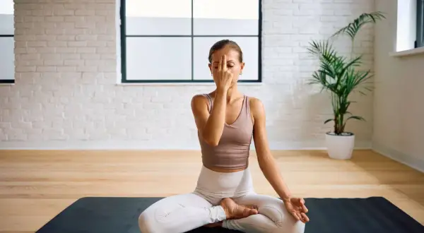 Frau praktiziert Pranayama Atmung im Lotussitz auf einer Yogamatte. Sie hält ihren Finger vor die Nase, um abwechselnd durch die Nasenlöcher zu atmen.