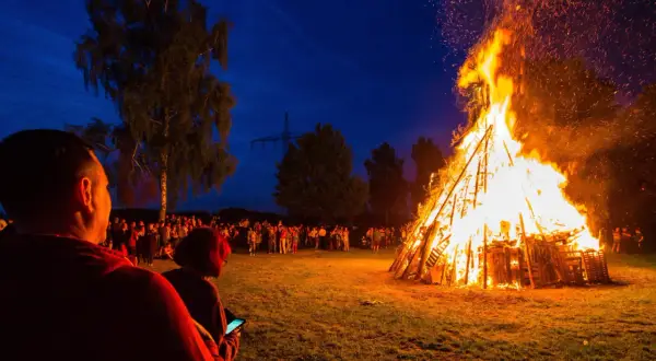 Menschen versammeln sich, um die Sommersonnenwende zu feiern. Ein riesiger Holzstapel brennt lichterloh im Abendhimmel, Funken steigen auf.