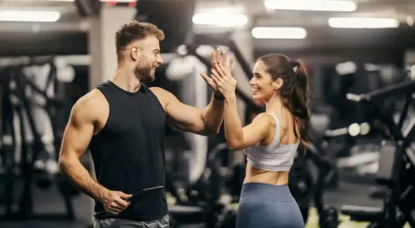 Mann und Frau geben sich im Fitnessstudio einen High Five. Er hält ein Tablet, vielleicht für seine persönliche Coach Ausbildung.