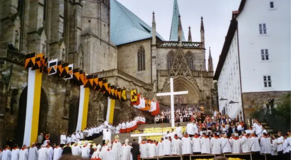 Große Menschenmenge vor einer Kathedrale, die an einem Hochamt teilnimmt. Fahnen und Girlanden schmücken die Fassade.