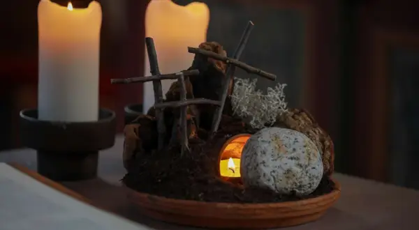 Grab und Kerzenlicht symbolisieren die **heilige Woche**. Ein Stein liegt vor dem Eingang einer Höhle, aus der ein Licht scheint.