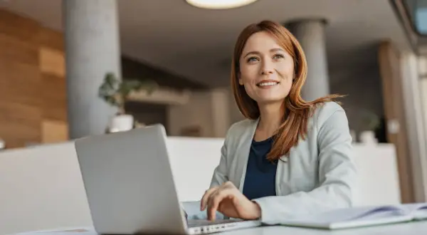 Eine Frau mit roten Haaren arbeitet an ihrem Laptop. Sie lächelt und blickt nach oben, als ob sie über ihre nächste Coaching-Sitzung nachdenkt. Ihr life coach job erfüllt sie.