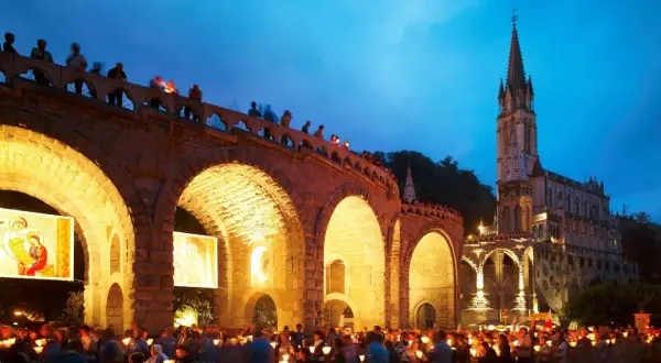 Eine Lichterprozession in Lourdes. Menschen halten Kerzen unter einem steinernen Bogen, während im Hintergrund eine Kirche beleuchtet ist.