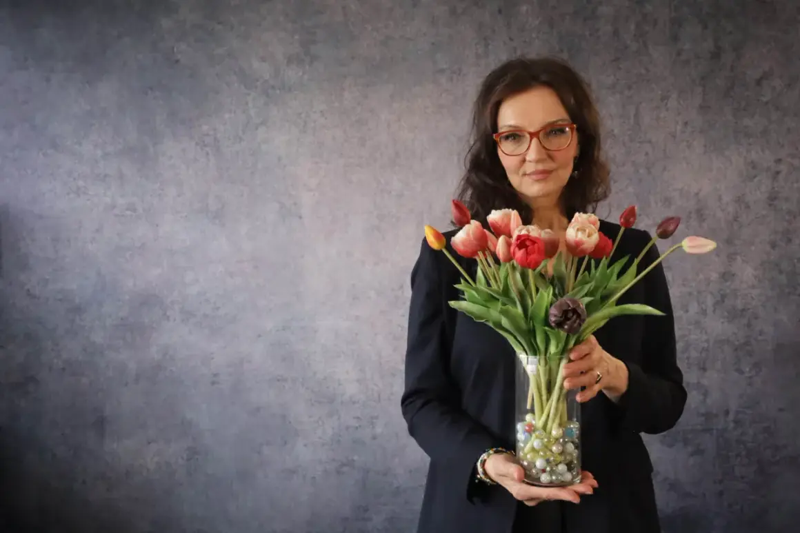 Eine energetische Person mit Brille hält einen Strauß bunter Tulpen in einem Glasvase.