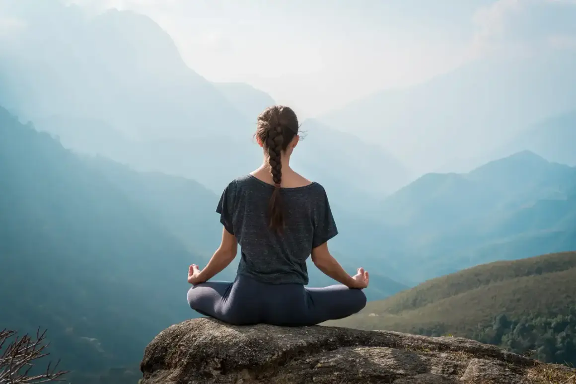 Frau in Hatha Yoga Pose auf einem Felsen sitzend, mit Blick auf eine neblige Berglandschaft.