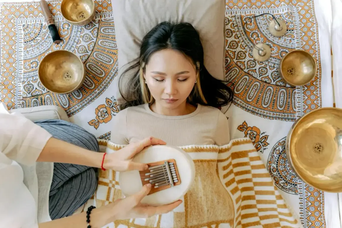 Eine Frau liegt entspannt, während eine andere Person ihr eine Kalimba über dem Bauch hält. Klangschalen und ein Stößel sind um sie herum arrangiert, was auf eine Klang Meditation hindeutet.